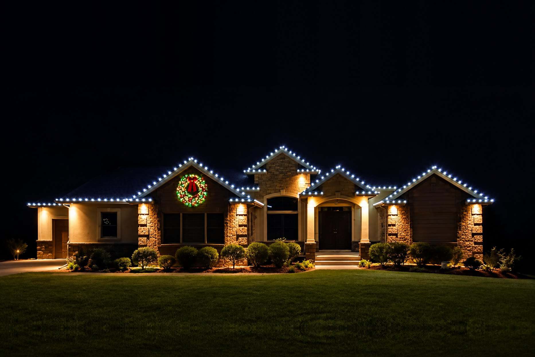 home-roofline-christmas-lights-multiple-peaks Ranch-style home at night with white lights along multiple rooflines and peaks and a lit wreath on the front gable
