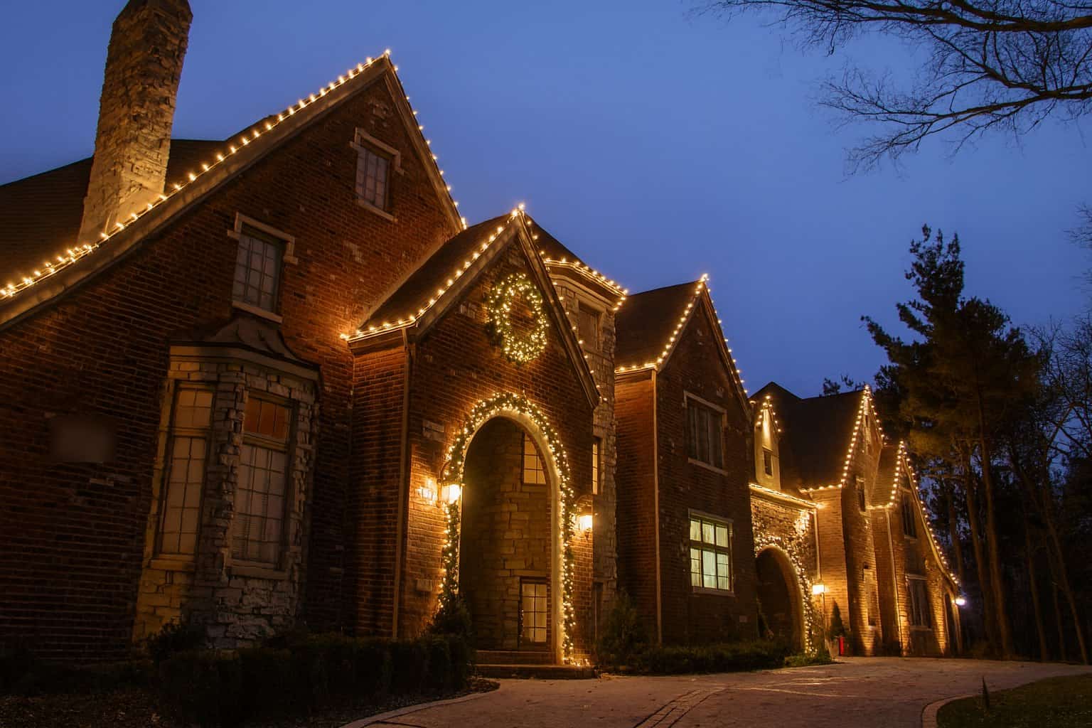 Brick home at dusk with warm white C9 lights along multiple rooflines and peaks, a lit wreath, and garland around the front archway
