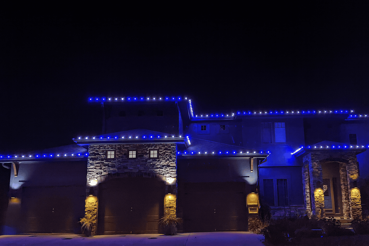 Christmas Light Design Ideas Two-story home at night with alternating blue and white C9 lights along multiple rooflines above a three-car garage and stone entry