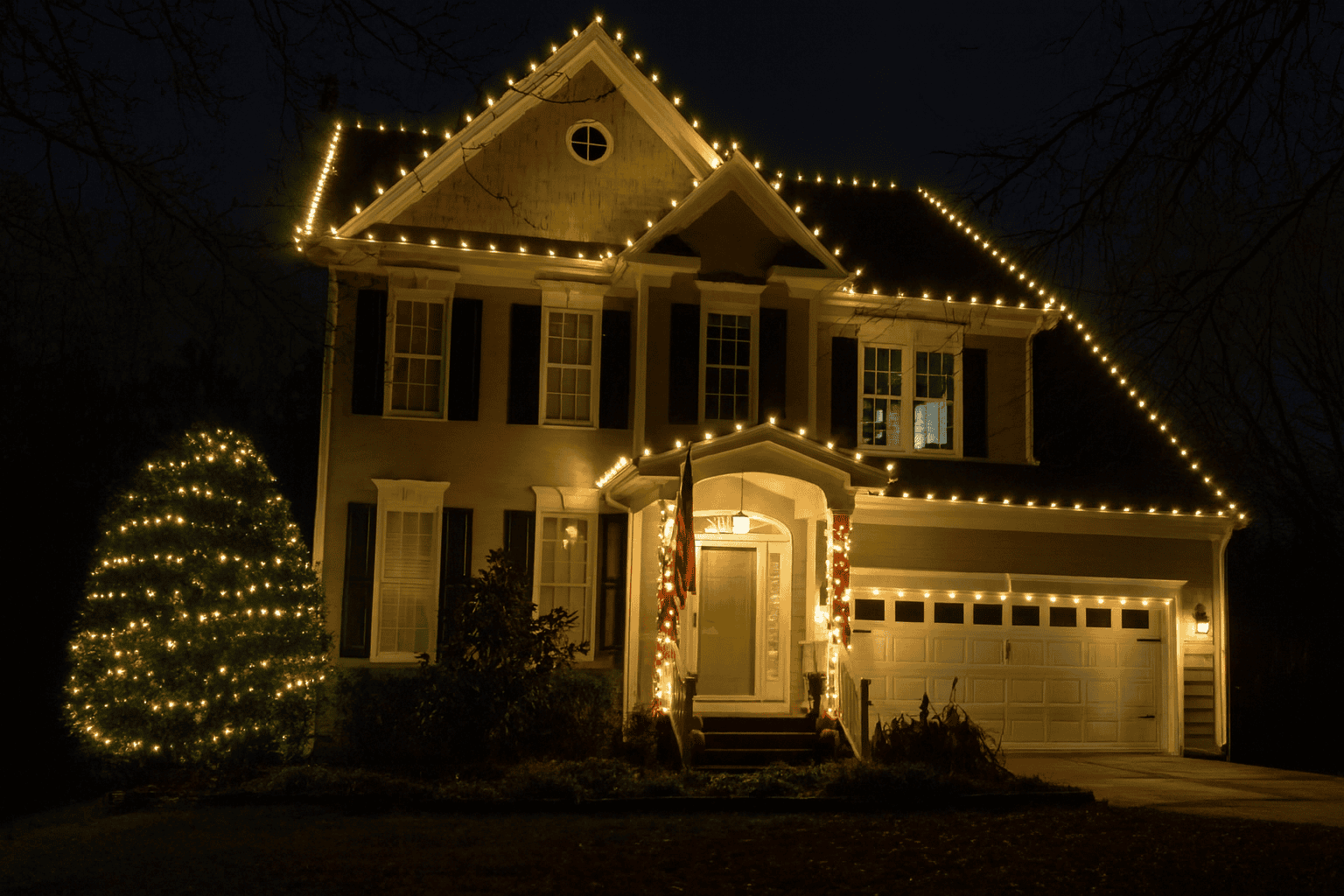 Two-story home at night with warm white C9 lights along every roofline and peak, a lit evergreen tree, and garland around the front entry